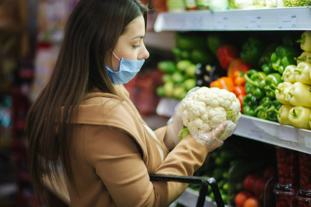 istock Happy woman in protective mask taking fresh vegetables while standing by groceries in supermarket. Beautiful young girl with food basket choosing food by stand with vegetables. Christmas purchases. Quarantine. Covid-19 1289159899