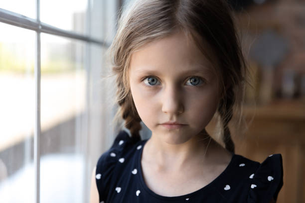 Portrait of serious little school age girl standing by window Close up portrait of pensive serious little school preschool age girl standing by window at home looking at camera with sad unhappy face having problem trouble thinking about bad relations in family autism photos stock pictures, royalty-free photos & images