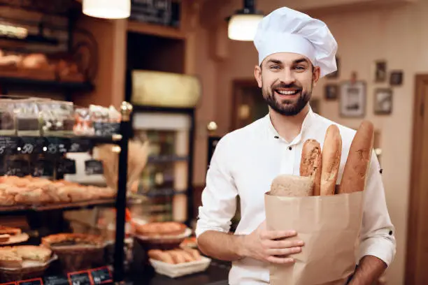 Male cook with a packet of fresh bread looks at the camera and smiles. Bearded man bakery worker with food in hands.