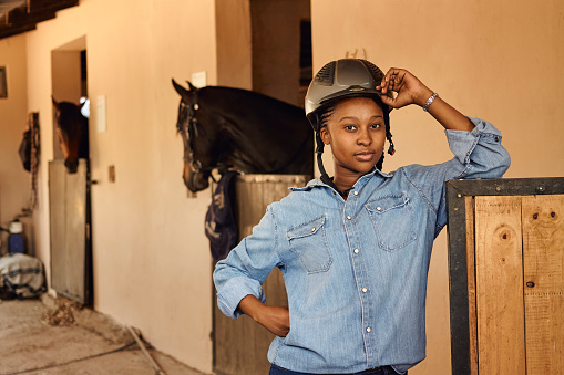 A young black African woman smiling blissfully looking into camera with her arm propped up on the stable door. She is wearing a blue button up shirt and a brown horse riding helmet while standing in the stables with a brown horse in the background