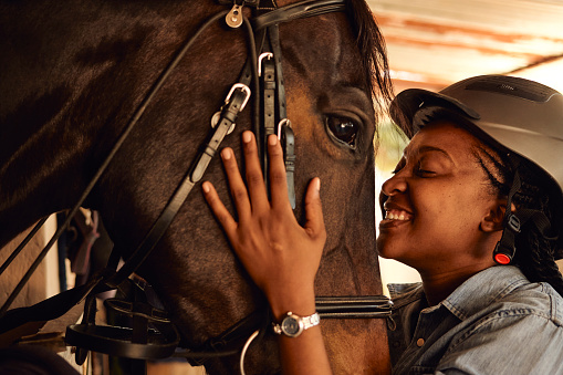 A young black African woman blissfully smiling big while holding her horse’s face embracing