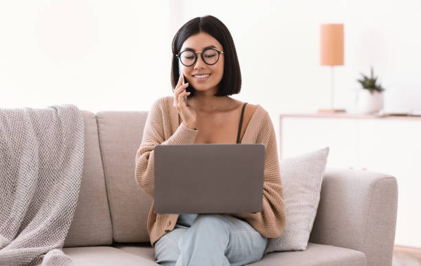 istock Portrait of smiling asian woman using pc talking on cellphone 1289177908