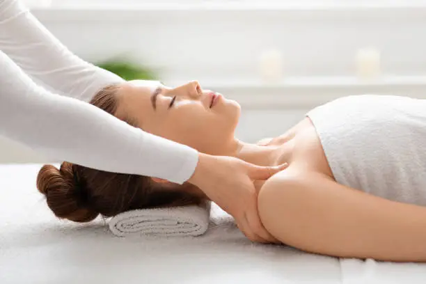 Side view of peaceful young lady having healing body massage from unrecognizable female therapist at modern newest spa. Spa attendant rubbing woman shoulders, stress relief concept