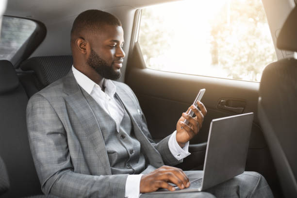 Handsome african businessman with laptop holding smartphone Handsome african american businessman with laptop holding smartphone while going by car on business trip, copy space. Black entrepreneur looking at mobile phone screen, using laptop black taxi stock pictures, royalty-free photos & images