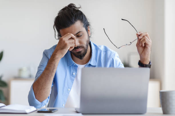 vermoeidheid van de ogen. jonge freelancer kerel masseren nosebridge, moe na het werken aan laptop - moe stockfoto's en -beelden