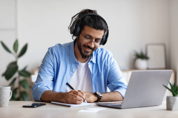 istock Western Guy Wearing Headphones Studying Online With Laptop At Home, Taking Notes 1289180594