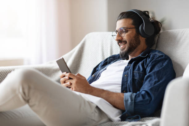 home leisure. young western guy in headphones relaxing with smartphone on couch - indian ethnicity audio imagens e fotografias de stock