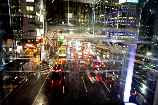 Urban landscape of Shibuya, Tokyo on a rainy night.\nA car is driving down an urban street on a rainy day.\nThe lights of the car are reflected in the wet road.
