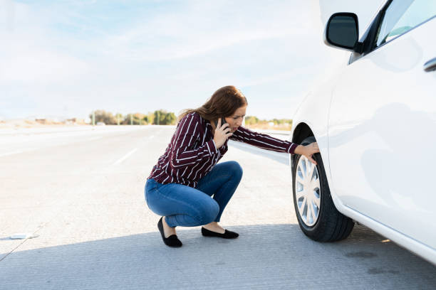 Woman in her 20s calling a tow truck on the phone because of a flat tire Worried young woman talking on the phone and checking her car flat tire on the side of the highway stranded stock pictures, royalty-free photos & images
