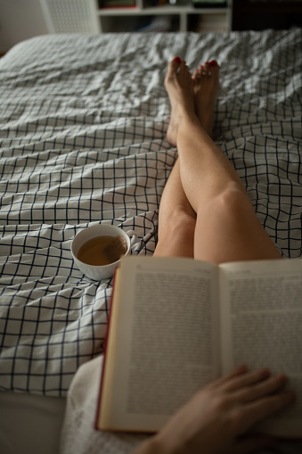 Mature woman holding cup of tea while sitting in bed in her bedroom and reading book. Photo taken from above. Photo taken as personal perspective view.\nPhoto on monitor display is available in my portfolio with ID #1135155166