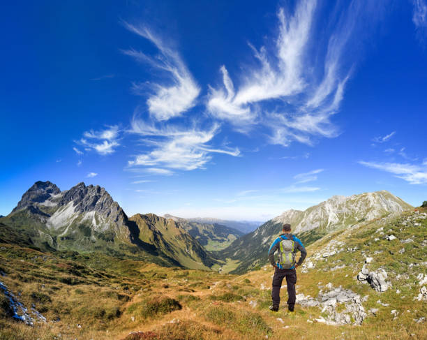 Hiker Man with Backpack enjoying great clear view in the mountain with beautiful clouds. Alps, Kleinwalsertal, Gemsteltal, Austria. Hiking Man with Backpack standing in Landscape enjoying clear view in the mountain with amazing clouds on deep blue sky. Alps, Kleinwalsertal, Gemsteltal, Austria. kleinwalsertal stock pictures, royalty-free photos & images
