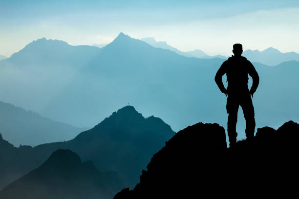 One Man reaching summit after climbing and hiking enjoying freedom and looking towards mountains silhouettes panorama during sunrise. stock photo