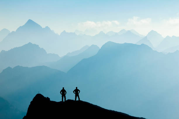 Two Men reaching summit after climbing and hiking enjoying freedom and looking towards mountains silhouettes panorama during sunrise. stock photo