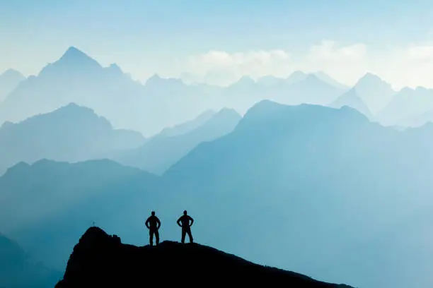 Two Happy winning successful silhouette hiker or climber at sunrise standing relaxed and are happy for having reached mountain top summit goal during alpine hiking and climbing adventure travel. Tirol, Austria and Allgaeu, Bavaria, Germany. Mountain Grosse Schlicke.
