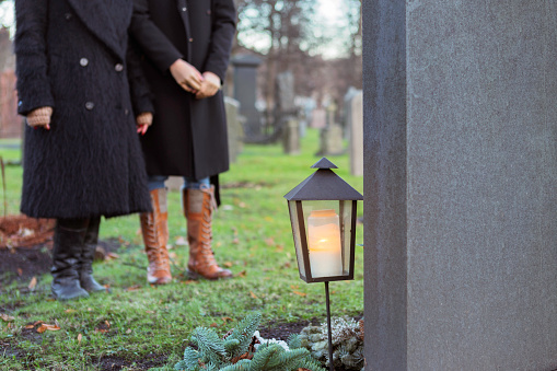 Two persons standing by a grave with a lit candle and lantern at the graveyard.