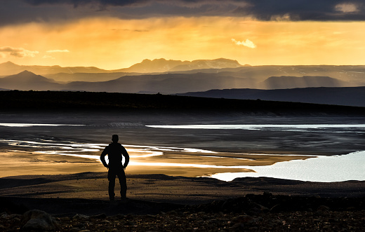 Hiking Man Silhouette watching orange sunlight sunset with fog over the mountains, river and lake. Amazing view of the landscape in Iceland.