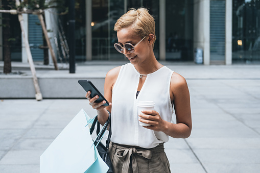 Blonde short hair woman standing at street in front of shopping mall. She is smiling, holding shopping bag and coffee while typing message on her smartphone