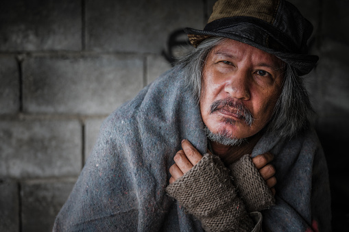 Close up and portrait of old homeless asian man gray hair and beard wear hat, gloves, sweater and blanket sitting cold with looking and seeking help on walkway street in city. Help and hope concept.