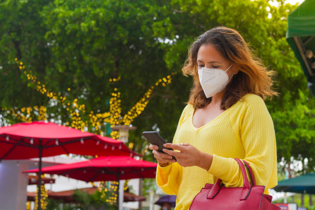 Young latin woman texting by cellphone waiting for a taxi in Miami Beach, Florida Young latin woman shopping and texting by cellphone waiting for a taxi at Lincoln Road in Miami Beach, Florida, USA. She's wearing a nice fashionable black eyewear, face mask, a yellow shirt, a red purse and jeans. 1 hotel south beach stock pictures, royalty-free photos & images