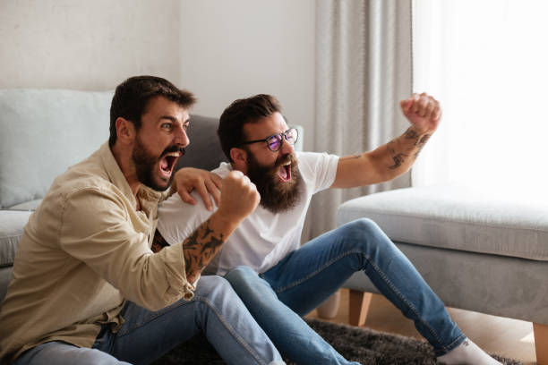 Two friends cheering, watching sport event on TV. Two young hipsters are watching football mach. It is final minute and score is even, but their favorite club is winning. They are cheering, smiling and hopping for their team to win. match sport stock pictures, royalty-free photos & images