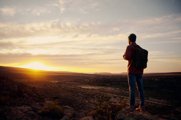 istock Low angle view of young male hiker with backpack standing on mountain top while hiking looking at sunrise 1289203409