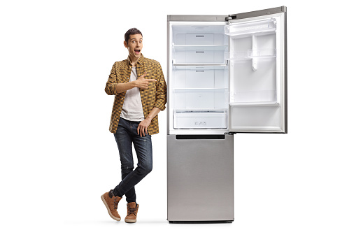 Full length portrait of a casual man leaning on a fridge and pointing isolated on white background