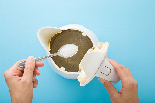 Young adult woman hand holding spoon with white powder of citric acid for descaling kettle. Closeup. Point of view shot. Light blue table background. Pastel color. Problem and solution. Top down view.