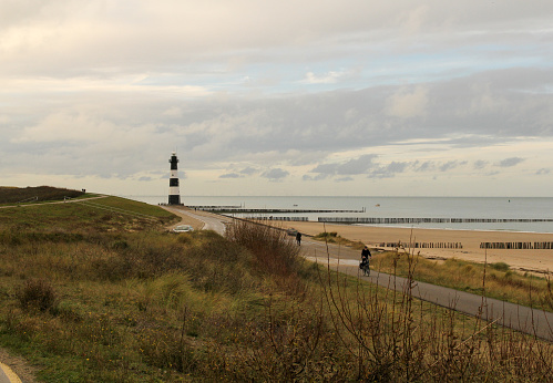 someone is cycling at a road in the dunes with a beautiful view at the sand beach and sea with lighthouse at the dutch coast in winter