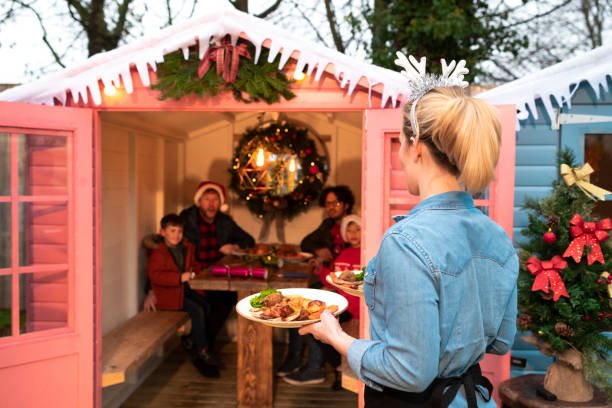 Christmas Dinner Outdoors An unrecognisable waitress serving Christmas dinner to a same sex parented family of four who are sitting in a shed which has a table and chairs inside and Christmas decorations. The family are wearing warm clothing and hats. gay dinner table stock pictures, royalty-free photos & images