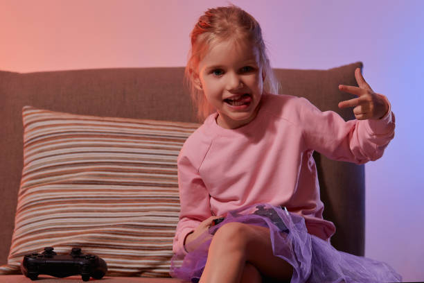 istock Cute little girl sit on couch and holding gamepad controller while playing video games at home, have fun in in living room with red blue neon light 1289210529