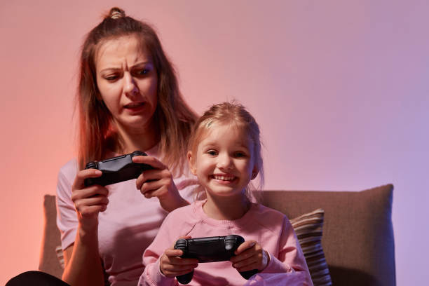 istock Excited young mother and cute little daughter sit on couch holding gamepad controller while playing video games at home together, have fun in in living room with red blue neon light 1289210542