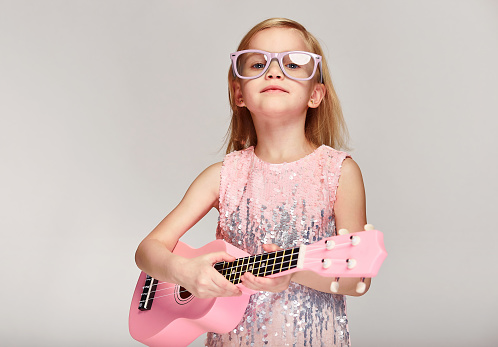 Rock and roll music concept.Cute caucasian little girl 4-5 year old in a bright pink dress and glasses are playing the ukulele on a gray studio background with copy space