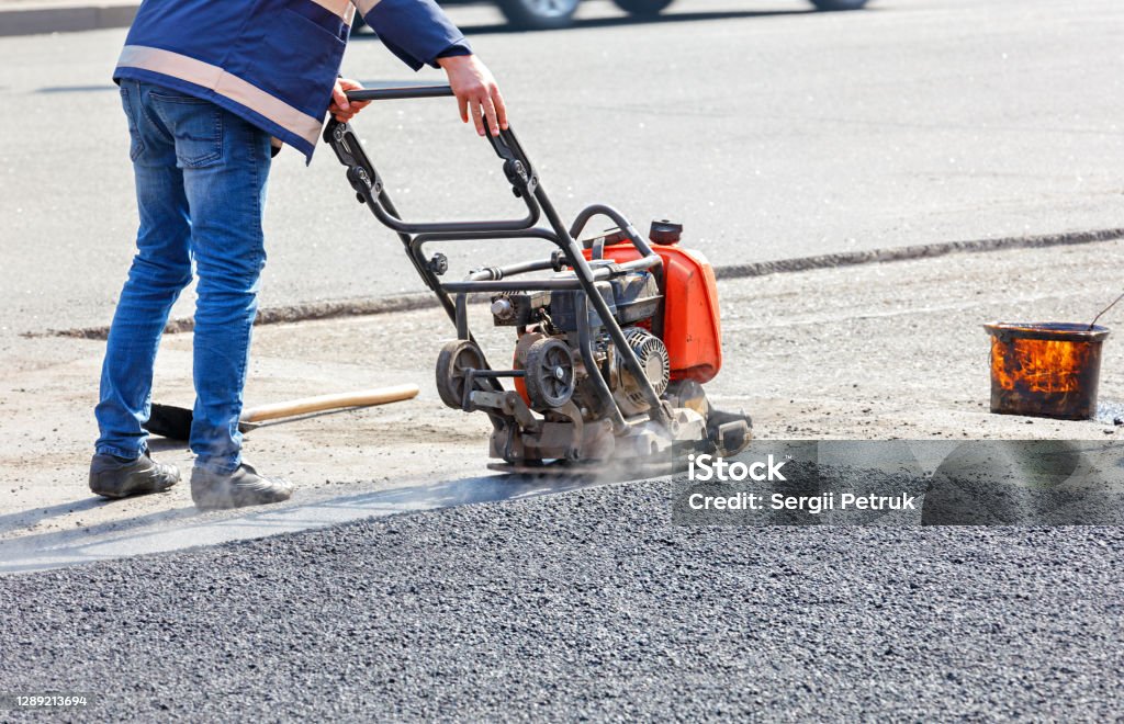 A road service worker repairs the roadway by compacting the asphalt with a plate petrol vibrator. A road service worker compacts the asphalt on a fenced road section of the roadway with a petrol vibration plate compactor. Activity Stock Photo