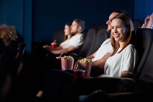 Selective focus of young girl laughing from weird situation, watching comedy in cinema. Side view of concentrated caucasian man and woman wearing white t shirts enjoying funny film.