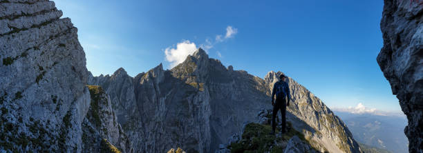 istock A man with a backpack standing in the shadow at the mountain peak and enjoying the view on higher, very steep peaks in front of him. Way to the Grimming in Austrian Alps. Freedom and exploration 1289216574