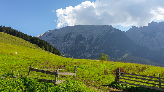 Distant view on Grimming, the highest self standing mountain in Alps, Austria. There is a lush green meadow in front and a wooden fence. Clear and bright day. Thicker clouds over the high Alpine peak