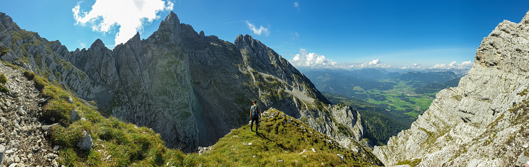 A man sitting at the edge of a mountain with a view on a vast valley. There are sharp mountains and high peaks around. The Alpine slopes are almost barren. Lush green valley. Bright day. Freedom.