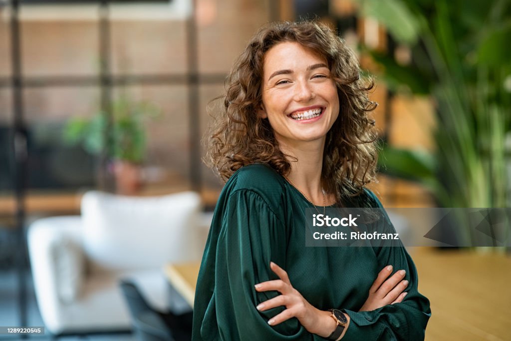 Beautiful woman smiling with crossed arms Portrait of young smiling woman looking at camera with crossed arms. Happy girl standing in creative office. Successful businesswoman standing in office with copy space. Women Stock Photo