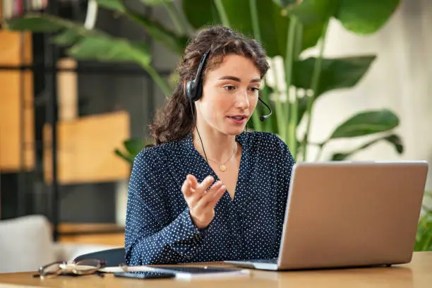 Confident young businesswoman wearing headphones and discussing project details with clients on video call. Skilled woman worker with headphones conducting online meeting with colleagues. Young casual girl talking to her colleagues about business strategy plan in video conference.