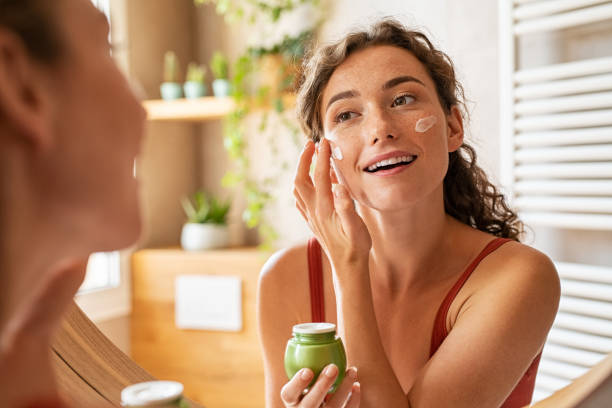Woman applying moisturiser on face during morning routine Woman caring of her beautiful skin face standing near mirror in the bathroom. Young woman applying moisturizing cream on her face during morning routine. Smiling natural girl holding little green jar of ecological cosmetic cream. beauty product stock pictures, royalty-free photos & images