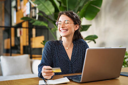Young happy woman doing shopping online with her laptop at home. Portrait of excited woman holding credit card and buy on an e-commerce site with copy space. Beautiful laughing girl paying online bills using debit card and computer.