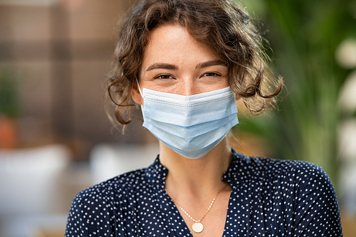 Portrait of happy young woman wearing face medical mask indoor. Hopeful girl with protective face mask looking at camera. Smiling woman wearing safety protective mask to fight against covid-19 pandemic.