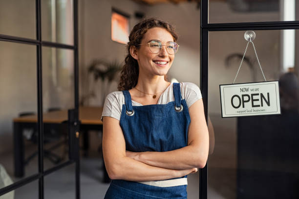 Small business owner standing at cafe entrance Portrait of happy waitress standing at restaurant entrance while looking outdoor. Portrait of young business woman attend new customers in her coffee shop. Smiling small business owner showing open sign in her shop while leaning against the door. small business owner stock pictures, royalty-free photos & images
