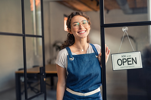 Portrait of positive business woman in apron standing at cafeteria door entrance. Cheerful young waitress in blue apron standing at glass door with open signboard and looking at camera. Excited small business owner standing at entrance of newly opened restaurant with open sign board.