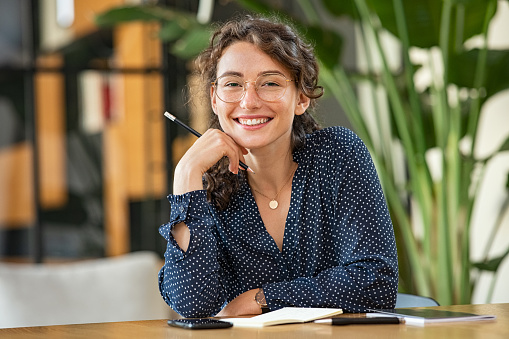 Portrait of smiling woman wearing spectacles and holding pencil while sitting at desk. Business woman taking notes in diary and looking at camera. University girl with eyeglasses sitting on table at library.