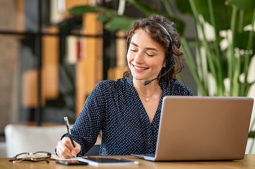 Successful young business woman working on laptop with headphones in a call center as a consultant. Happy businesswoman with headset translating and writing notes by listening to audio course while studying on laptop. Happy university student girl attending online class.
