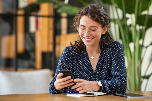 Young smiling business woman  sending text message and sms with smartphone at desk. Casual woman wearing eyeglasses and using mobile phone to check email at work with copy space. Cheerful student girl sitting at table and messaging online with cellphone.