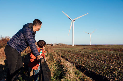An Asian father and son leaning against a fence in a field together, they are looking at the wind turbines.