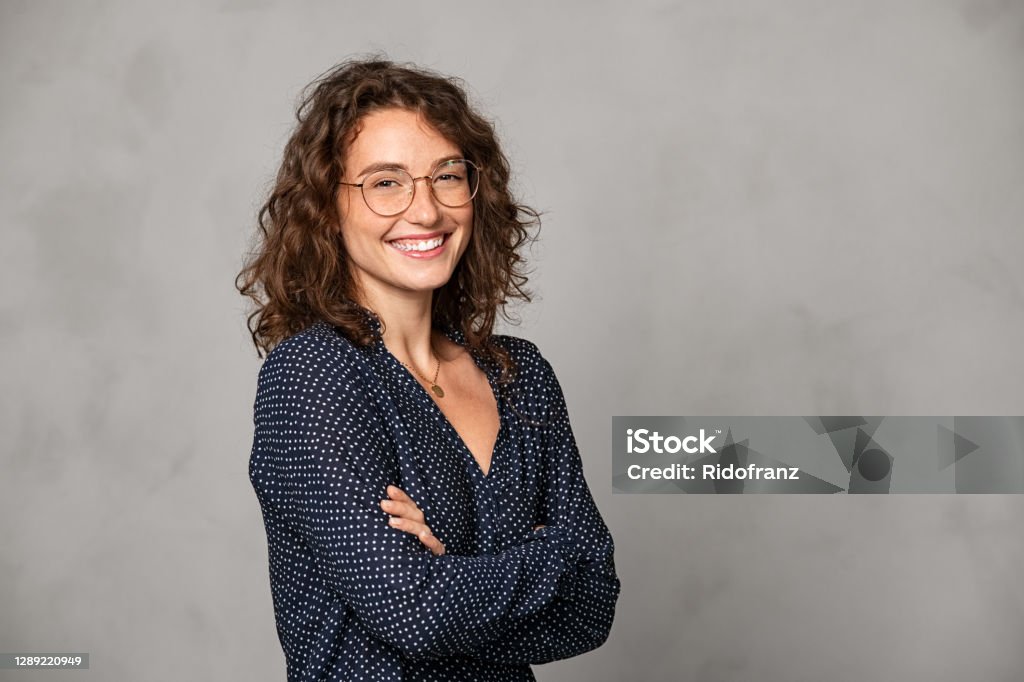 Successful smiling woman wearing eyeglasses on grey wall Confident young woman wearing eyeglasses and standing with folded arms on gray wall. Portrait of smiling businesswoman with arms crossed isolated against grey background with copy space. Proud university student girl with specs looking at camera. Women Stock Photo