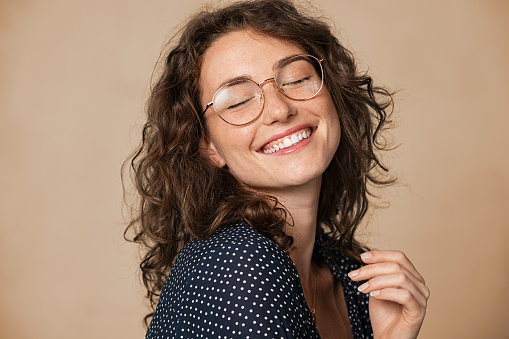 Casual cheerful woman with eyeglasses smiling at camera on cream background. Close up of happy young woman laughing with eyeglasses. Beautiful natural girl having fun with closed eyes showing a big grin.